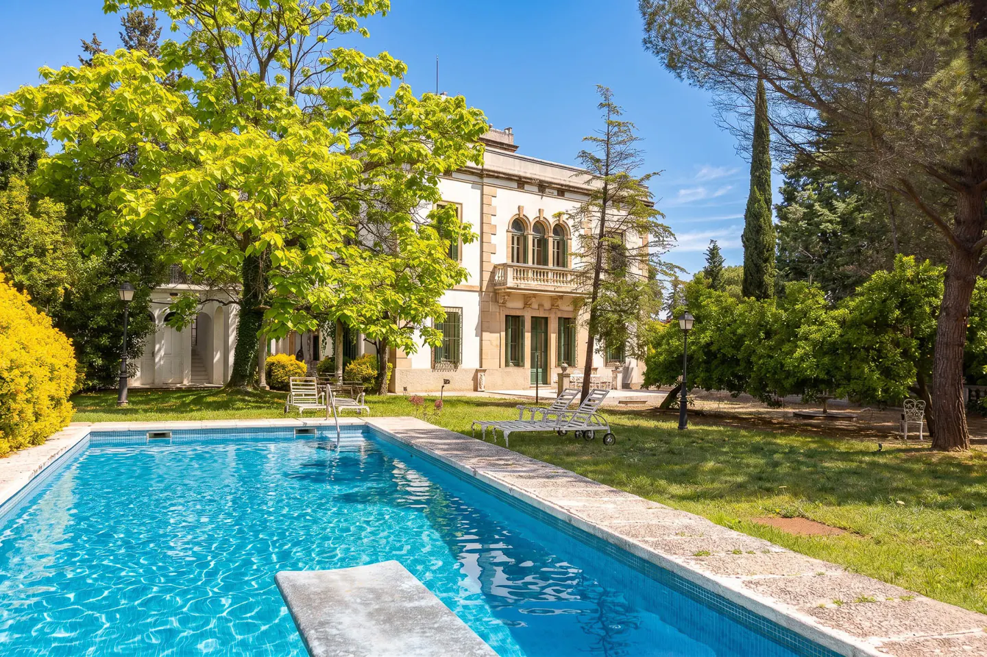 Exterior view of a large, white house with a pool in the foreground, surrounded by green trees and grass.