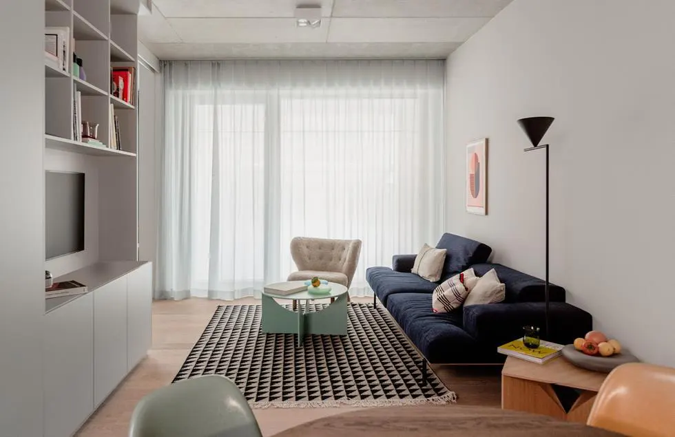 A modern living room with a blue sofa, patterned rug, and light gray walls. A bookshelf and TV are on the left, with a window and sheer curtains in the background.