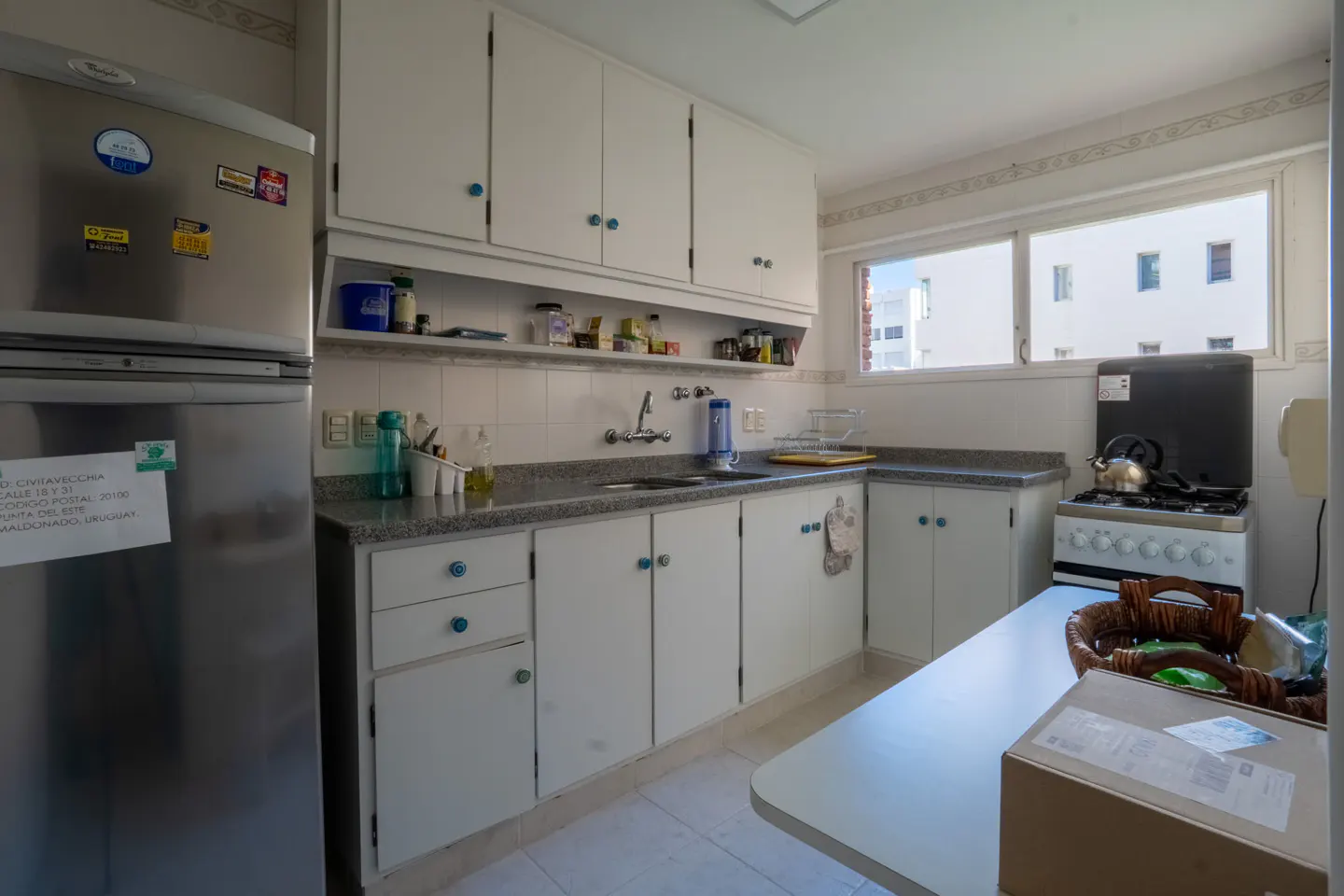 Bright kitchen with white cabinets, gray countertops, and stainless steel fridge. A window shows a white building outside.