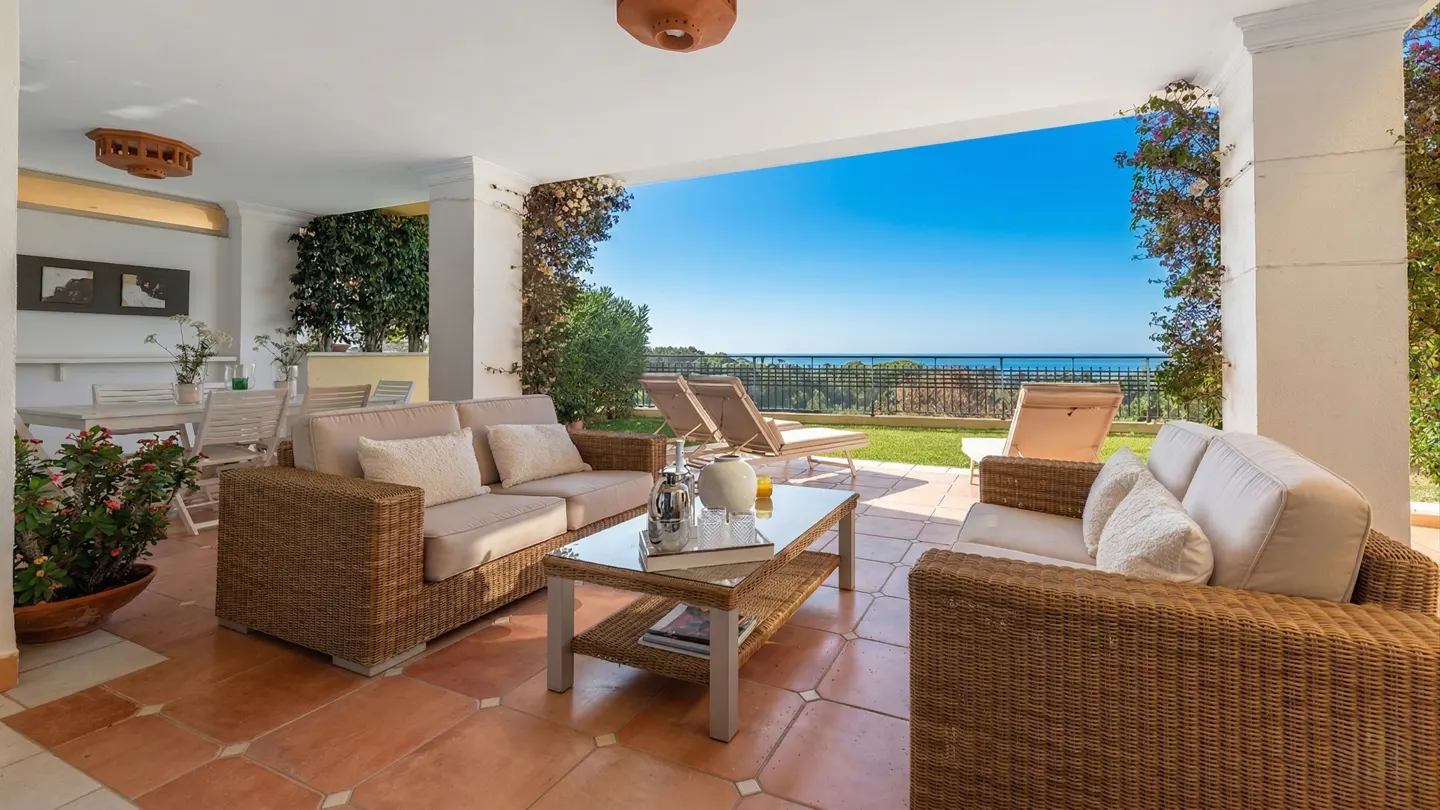 Covered patio with wicker furniture, a glass table, and a view of the ocean and blue sky.