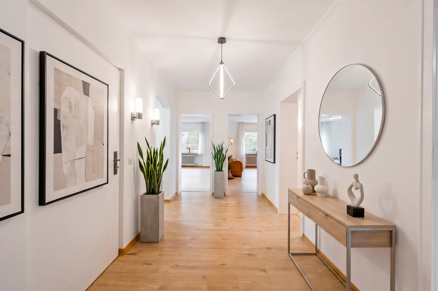 Bright hallway with wood floors, white walls, and modern decor. Artwork, plants, a console table with a round mirror, and a diamond-shaped pendant light are visible.