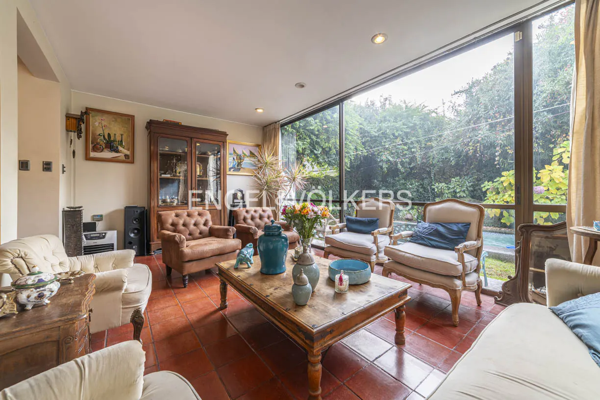 Living room with terracotta floors, leather and upholstered armchairs, a wooden coffee table, and large garden view windows.