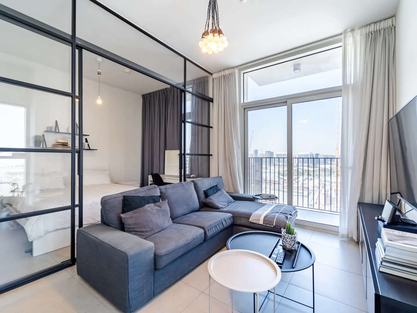 Modern living room with a grey sectional sofa, glass partition wall, and balcony view of the city skyline.