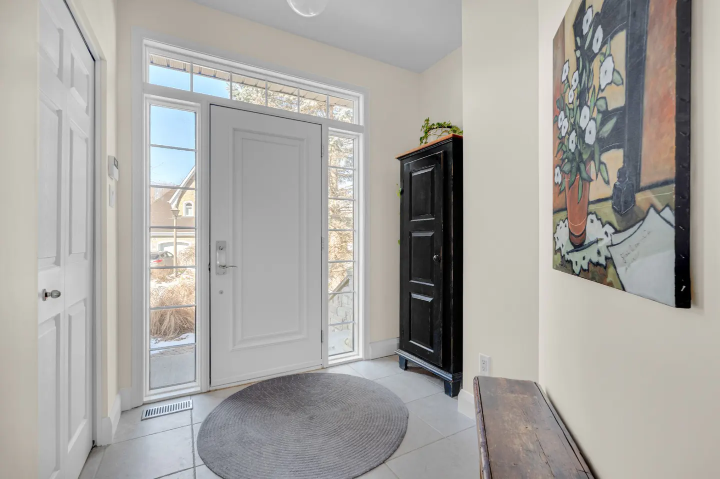 Foyer with white door, sidelights, and transom window. A black cabinet, wooden bench, and floral painting decorate the light beige walls.