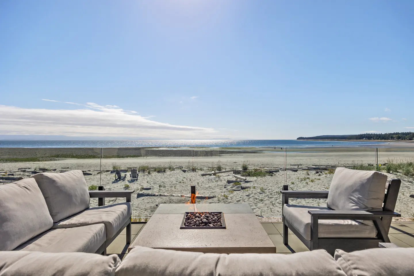 Outdoor patio with a fire pit, sofa, and chair overlooking a sandy beach and ocean under a blue sky.