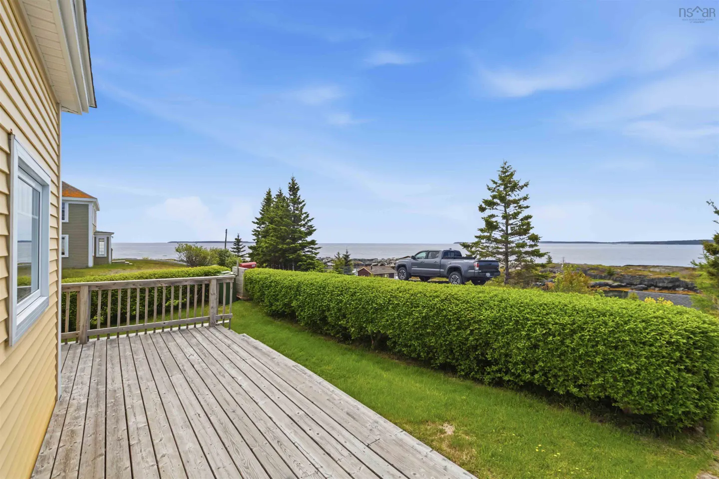 Wooden deck overlooking a green hedge, a parked grey pickup truck, and a scenic ocean view under a clear blue sky.