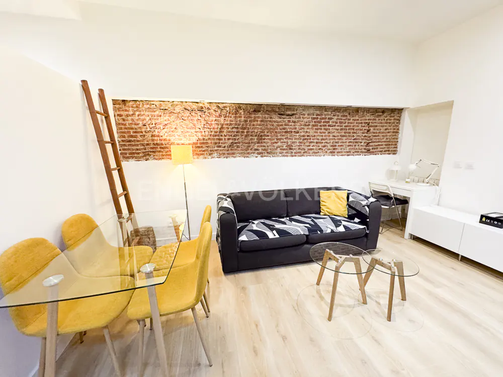 Bright living space with yellow chairs, glass table, black sofa, and exposed brick wall. A wooden ladder leans against the wall.