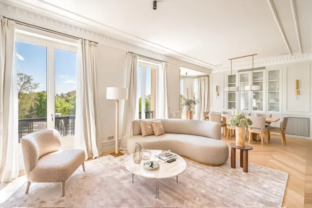 Bright, airy living room with beige curved sofa, chair, and marble coffee table on a light rug. Dining area visible in the background.