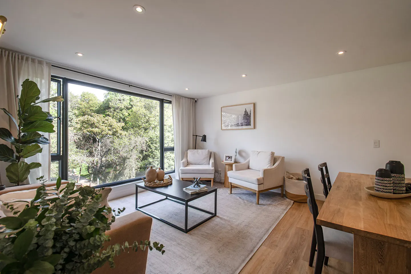 Bright living room with large window view of trees. Two white armchairs face a black coffee table on a neutral rug. Wood dining table with black chairs.