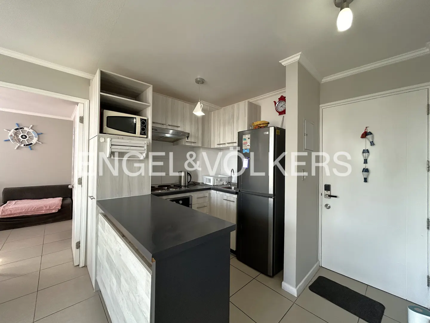 A kitchen with gray cabinets, a black countertop, and a black refrigerator. A living room is visible through a doorway.