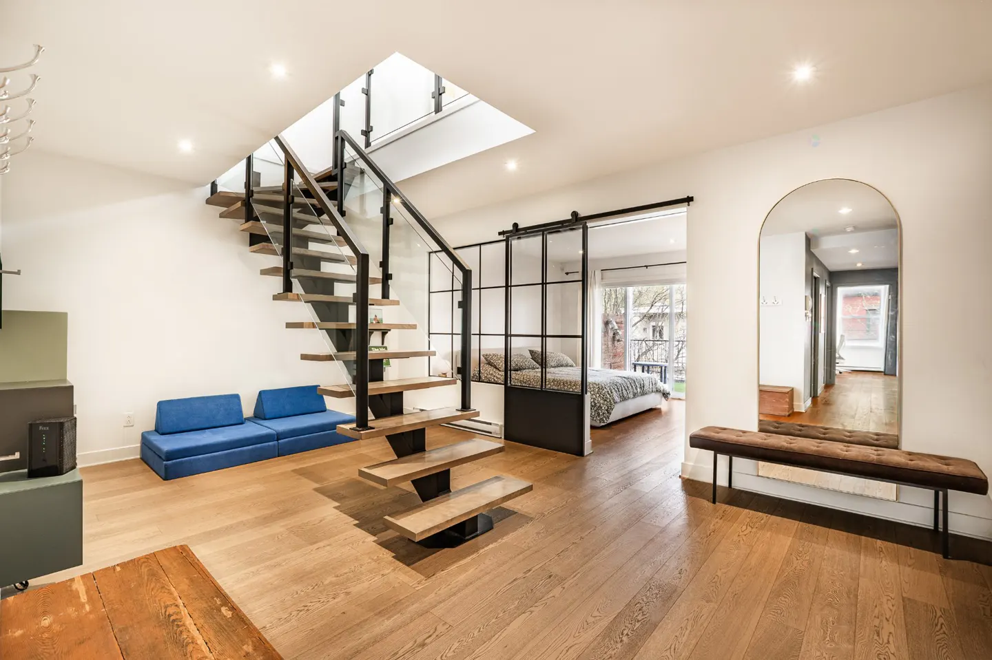 Bright, modern interior with wood floors, floating stairs, blue sofa, and sliding glass door to bedroom. A large mirror reflects a hallway.