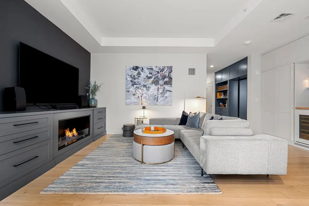 Living room with gray sectional sofa, blue rug, and fireplace under a large TV. Abstract art hangs on the wall.