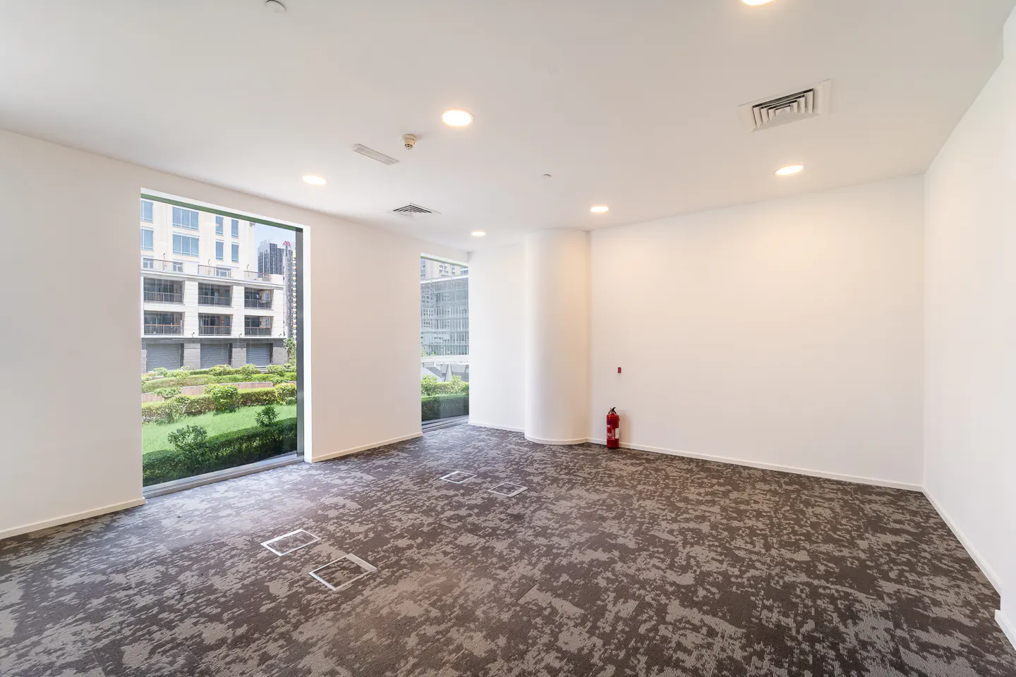Bright, empty office space with white walls, grey patterned carpet, and large windows overlooking a green landscape and buildings. A red fire extinguisher is visible.