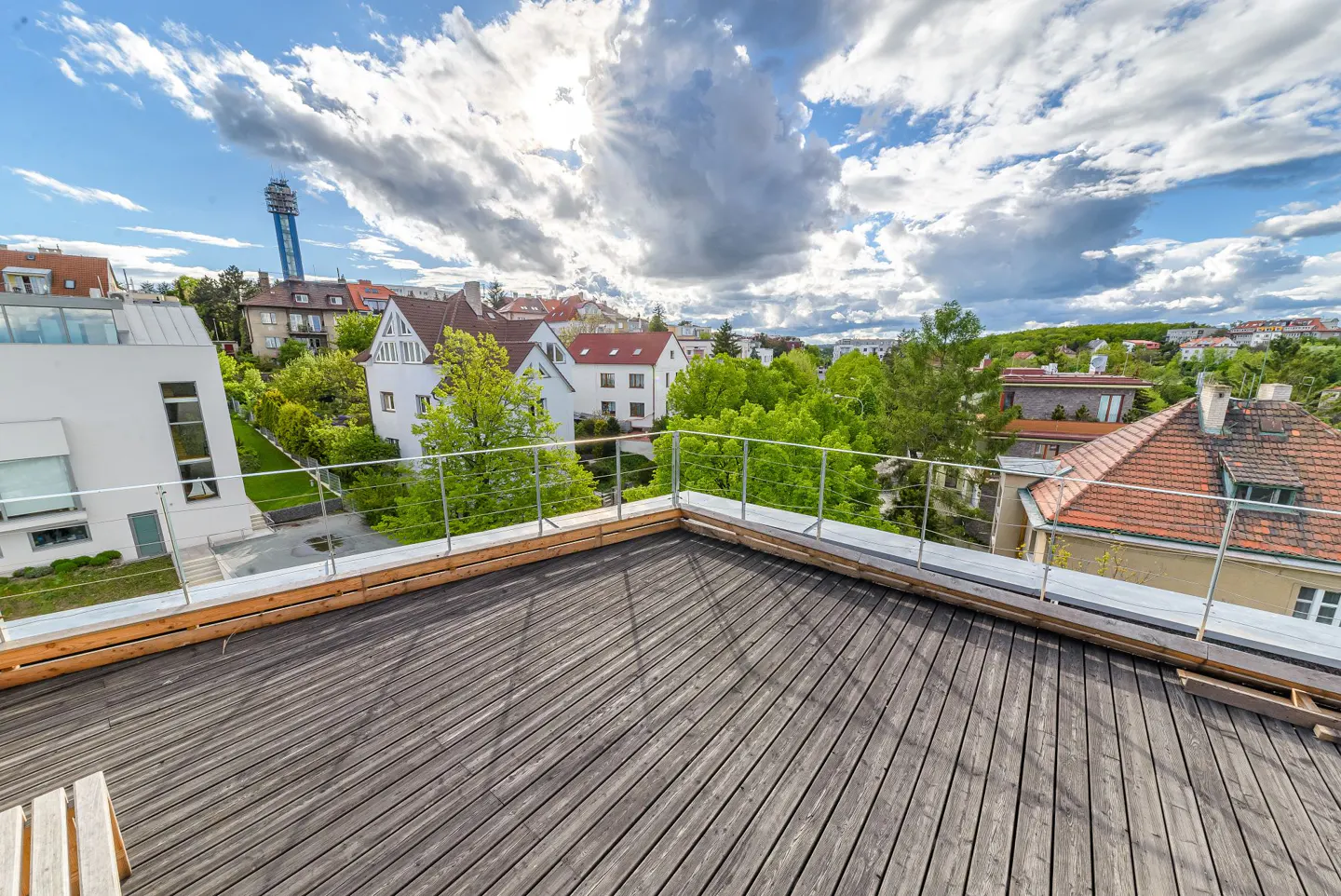 Rooftop terrace with wooden decking and metal railings overlooking a neighborhood with houses, trees, and a cloudy sky.