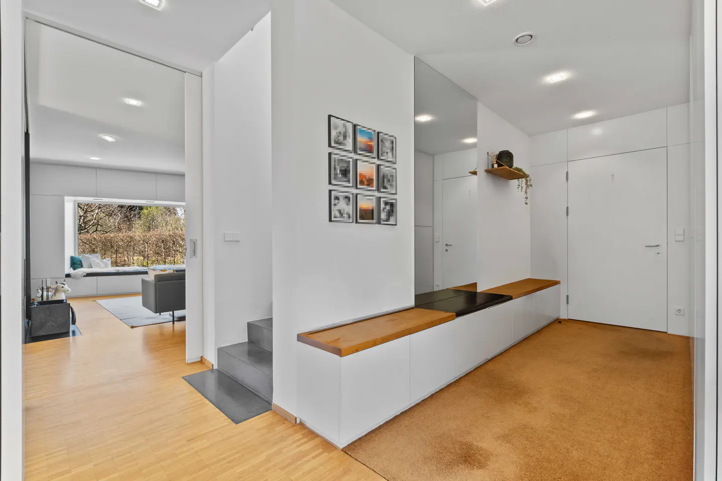 Bright entryway with wood floors, white walls, and built-in bench seating. A mirror and framed photos decorate the wall. A glimpse of the living room is visible.