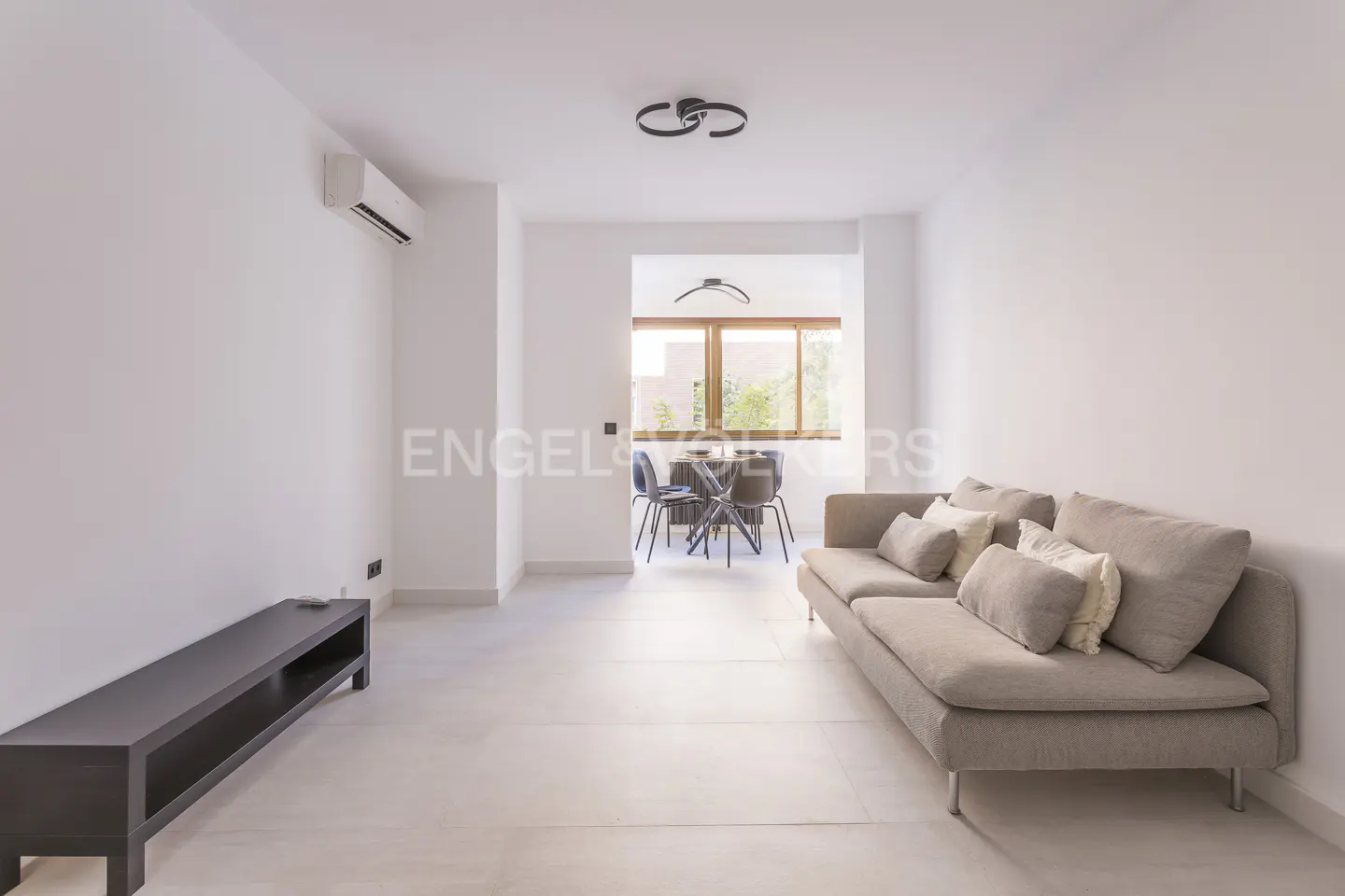 Bright, modern living room with white walls, a beige sofa, and a dark brown TV stand. A dining table is visible through an opening.