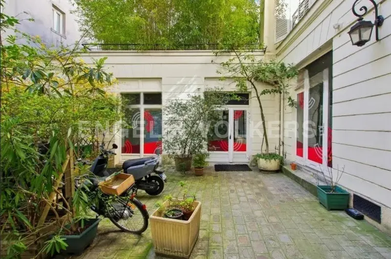 A courtyard with a scooter, bicycle, and plants in pots. The building is white with red accents and a bamboo garden on the roof.
