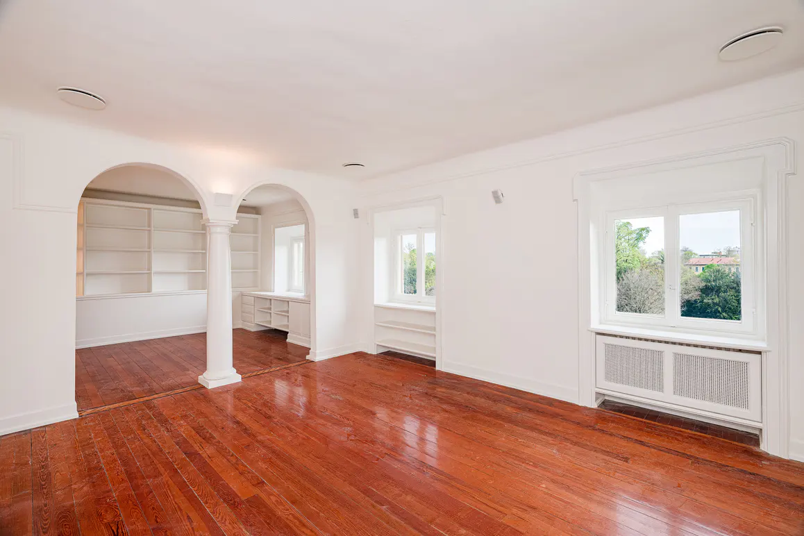 Bright, empty room with shiny wood floors, white walls, arched doorway to a built-in bookcase, and a window with a radiator.