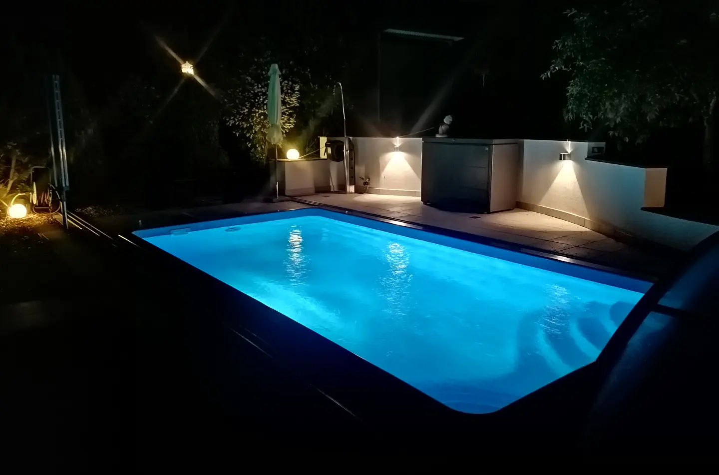 Night view of a rectangular pool with bright blue water, surrounded by dark greenery and soft lighting.
