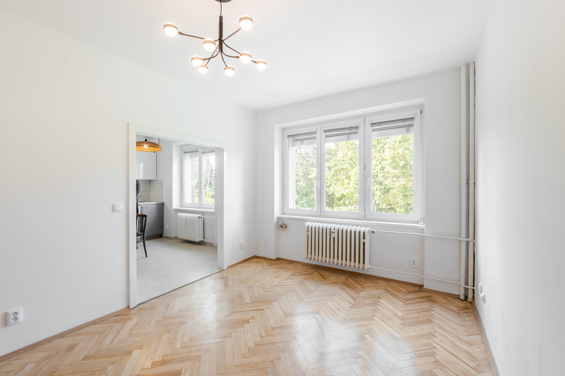 Bright, empty room with light wood herringbone floors, white walls, a modern chandelier, and a view of trees outside. [AI-generated text]