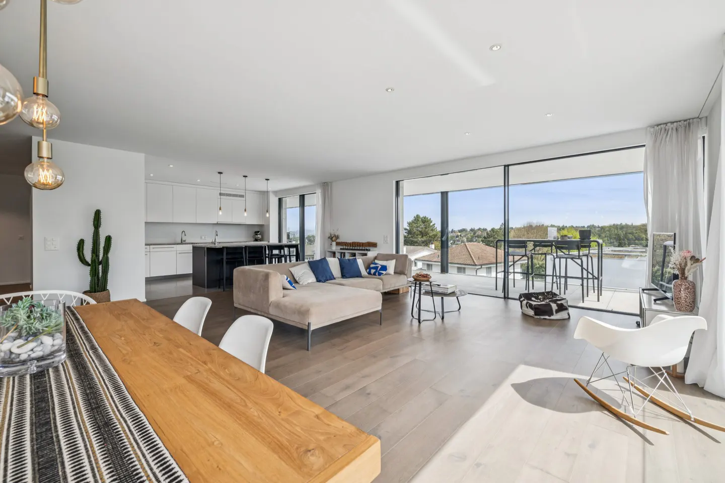 Bright, open-concept living space with a wood dining table, beige sectional sofa, and floor-to-ceiling windows overlooking a balcony.