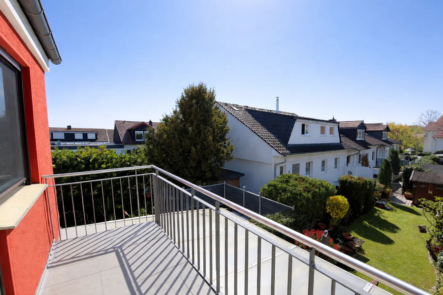 Balcony view of a neighborhood with houses, trees, and a green lawn on a sunny day. The balcony has a metal railing and gray tiles.