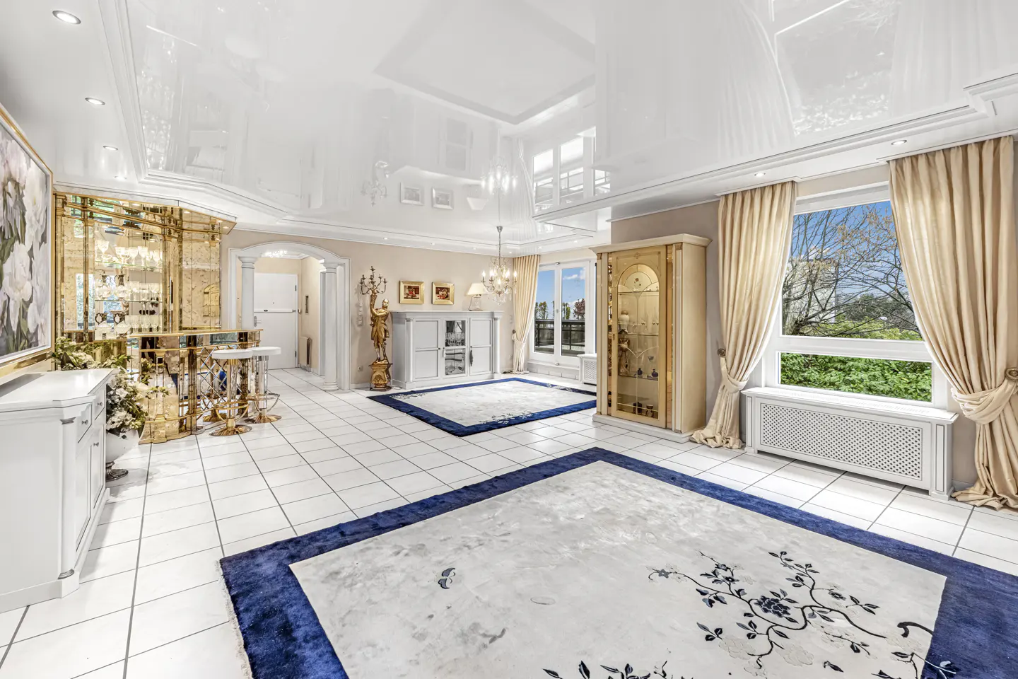 Opulent living room with white tile floors, gold bar, and blue-bordered rugs. Windows with beige curtains overlook greenery.