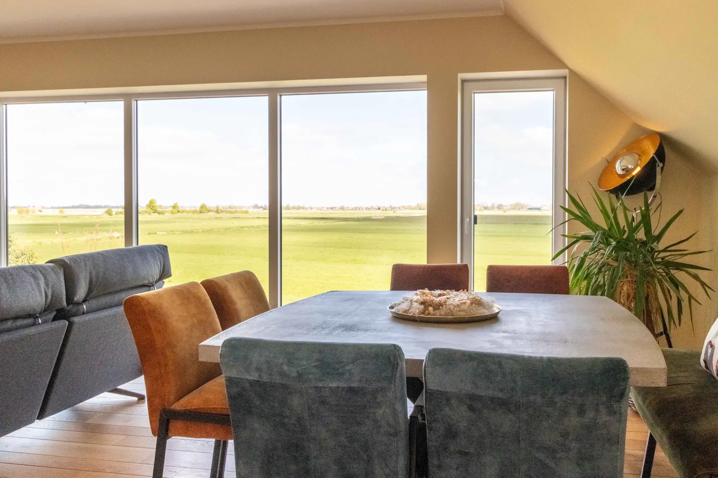 Bright dining room with large windows overlooking a green field. A gray table is surrounded by velvet chairs in blue, orange, and brown.
