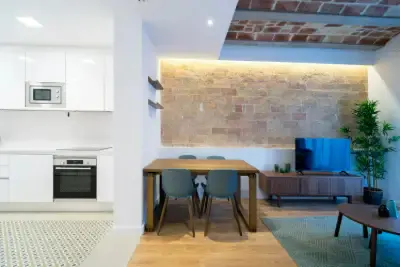 Open-concept apartment with white kitchen, wood dining table with blue chairs, brick wall, and living area with TV.