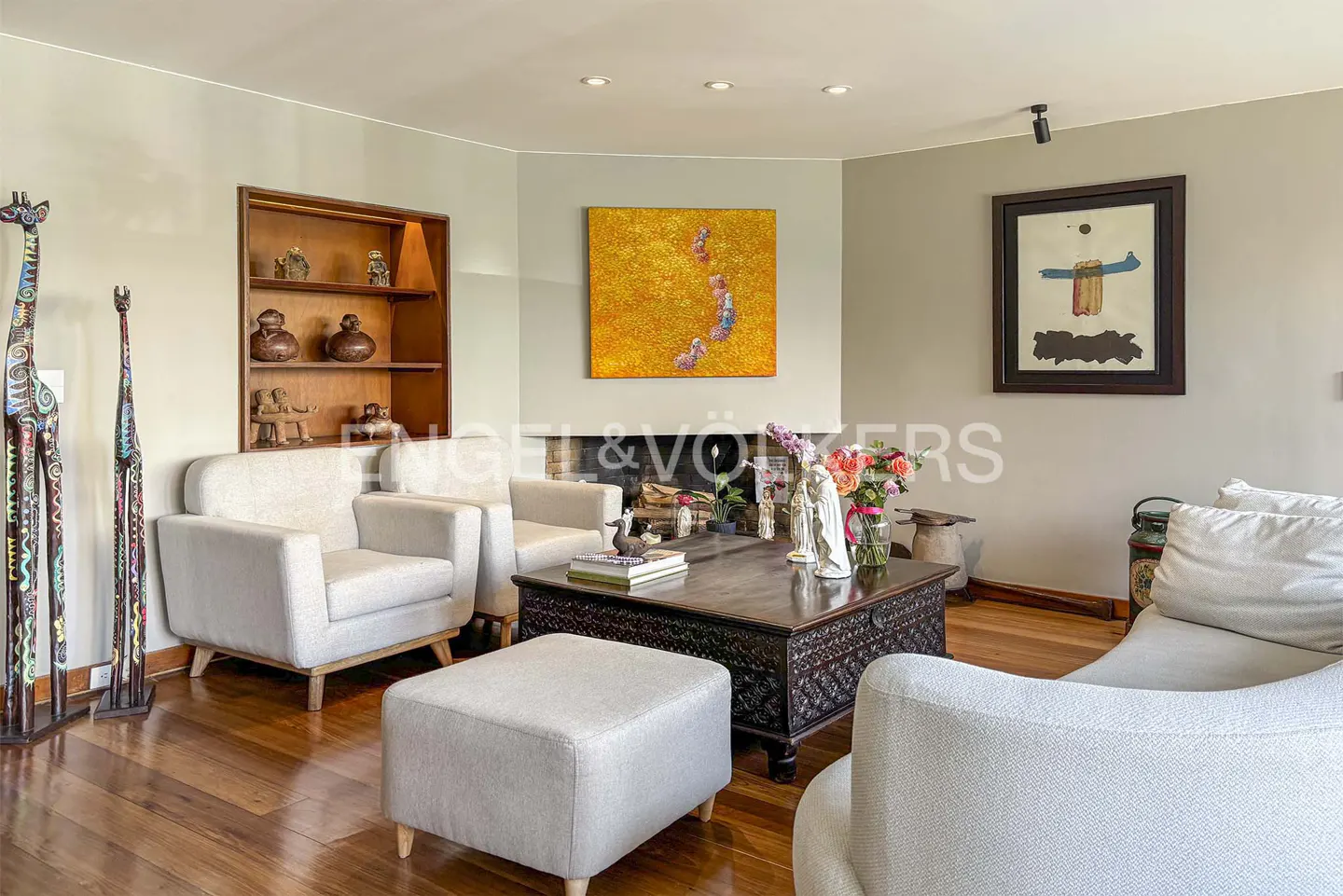 Living room with hardwood floors, light gray sofas, and a dark wood coffee table with flowers. Artwork hangs above the fireplace and on the wall.