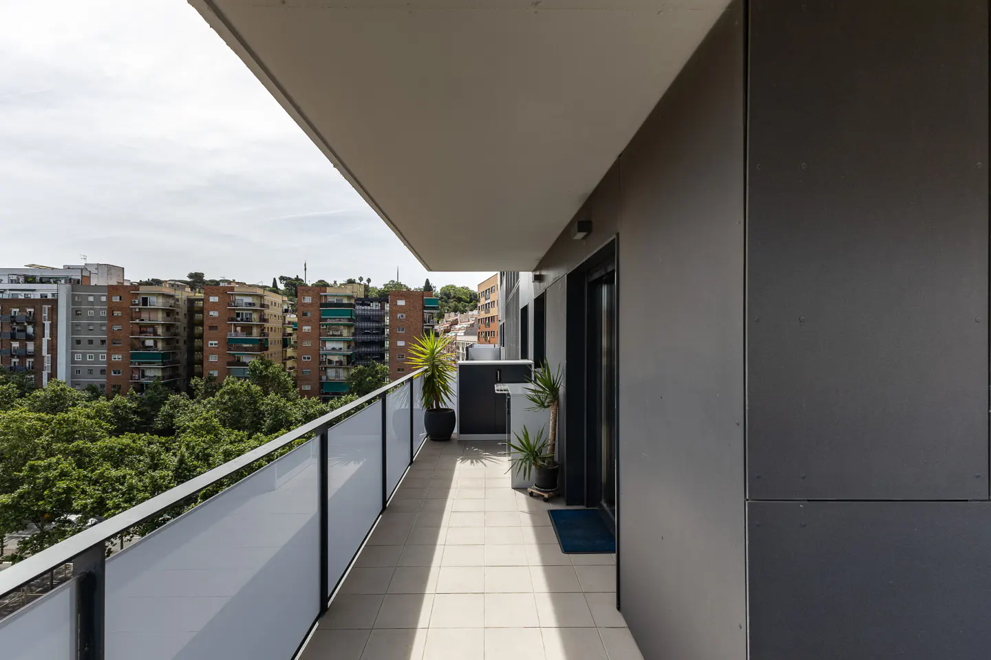 Modern apartment balcony with tiled floor, glass railing, potted plants, and a view of city buildings and trees.