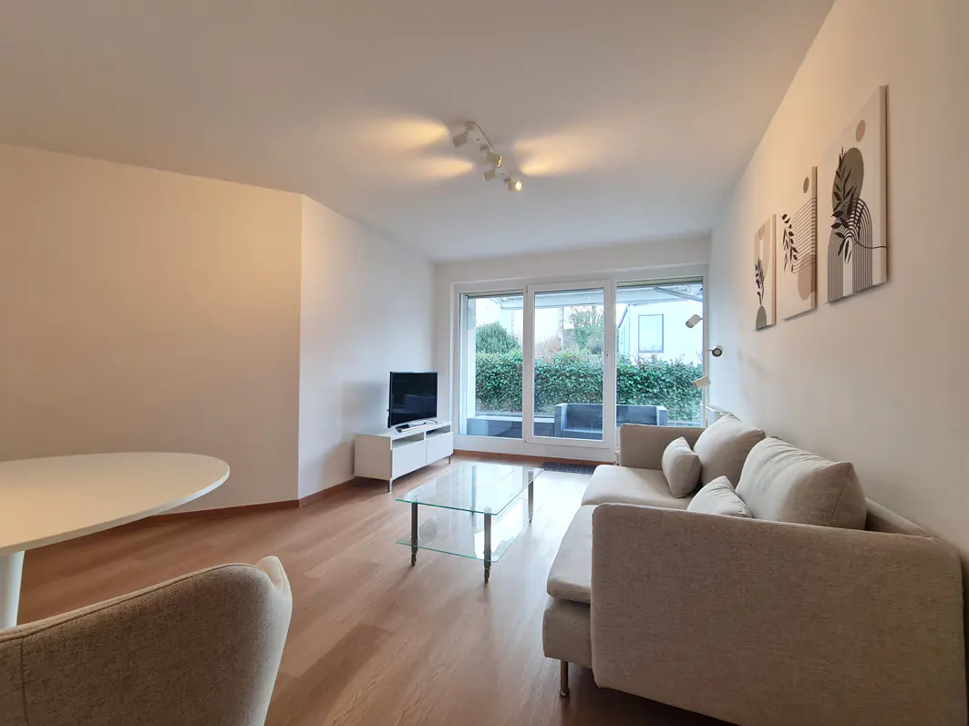 Bright living room with light wood floors, beige sofa, glass coffee table, TV stand, and abstract wall art.