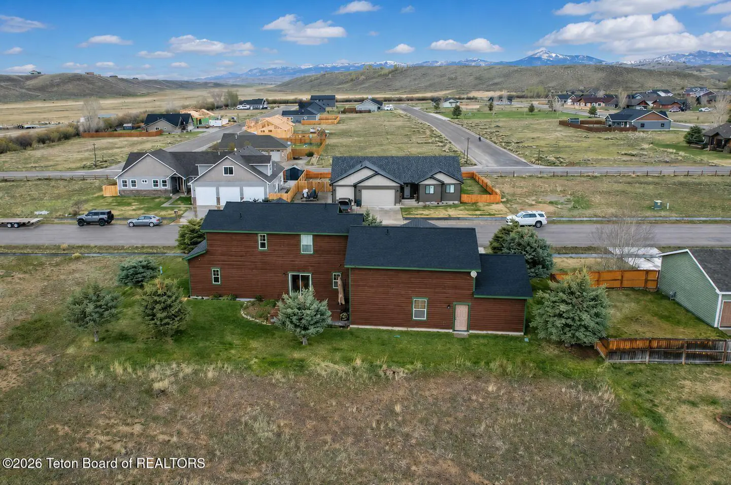 Aerial view of a brown two-story house with a black roof in a rural neighborhood with mountains in the background.