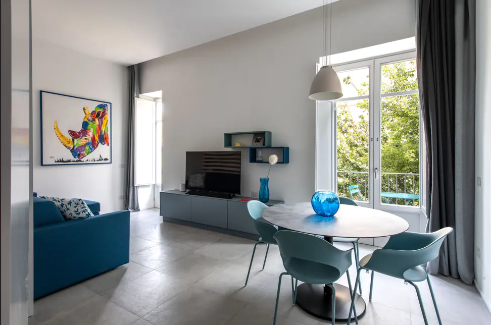 Bright living room with blue sofa, colorful rhino art, TV, and round table with teal chairs near a window with green trees outside.