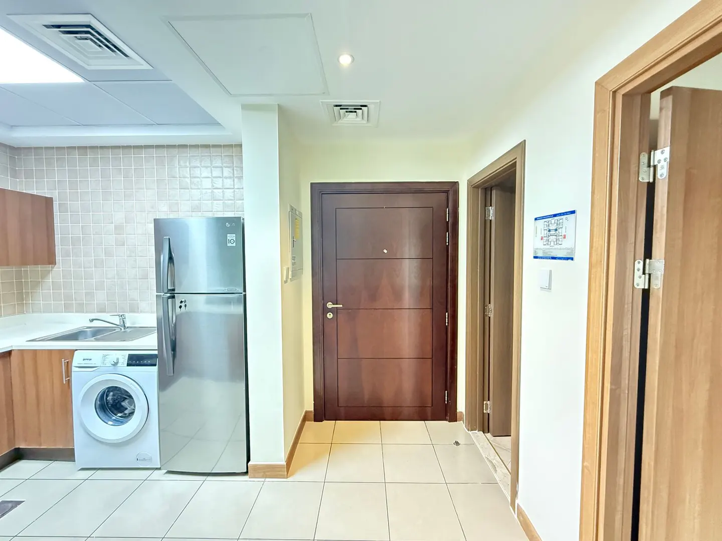 Interior view of a kitchen with stainless steel refrigerator, washing machine, sink, and brown cabinets. There are three doors in the hallway.