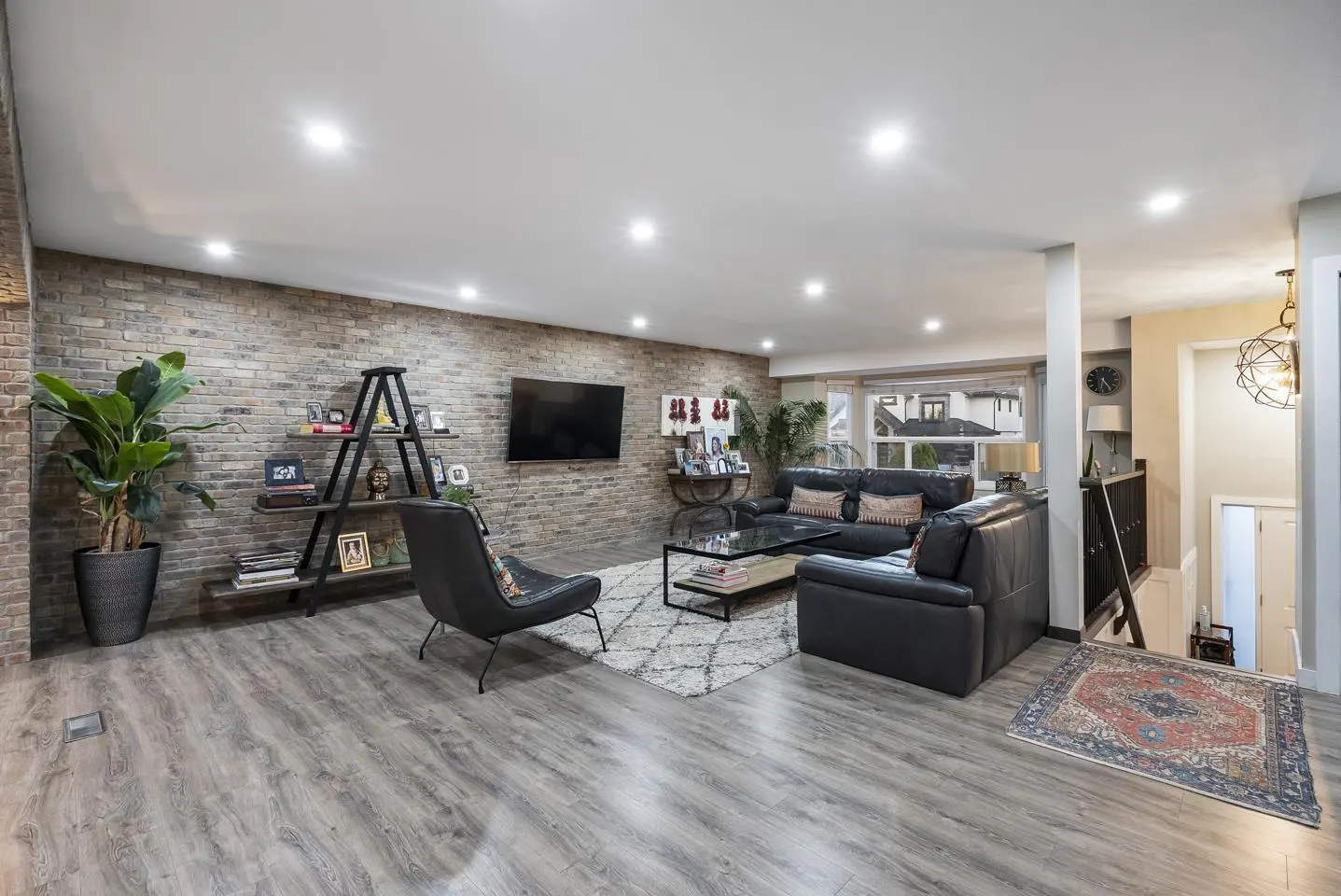 Living room with gray wood floors, brick wall, black leather sofa and chair, and a white rug. A staircase is visible on the right.