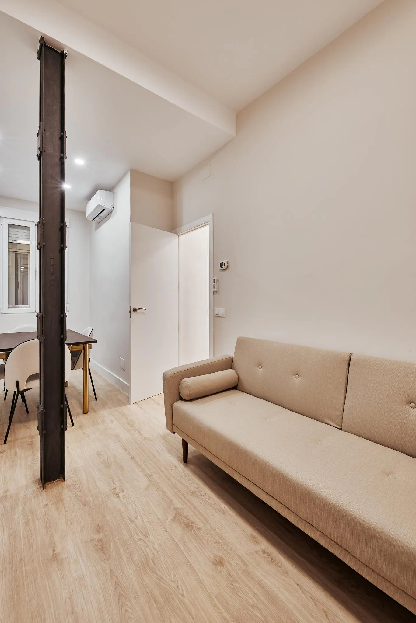 Living room with a beige sofa, light wood floors, and a dark metal support beam. A dining table and chairs are visible in the background.