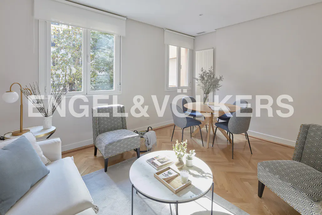 Bright living room with herringbone wood floors, white walls, and large windows. Features a sofa, chairs, round table, and dining set.