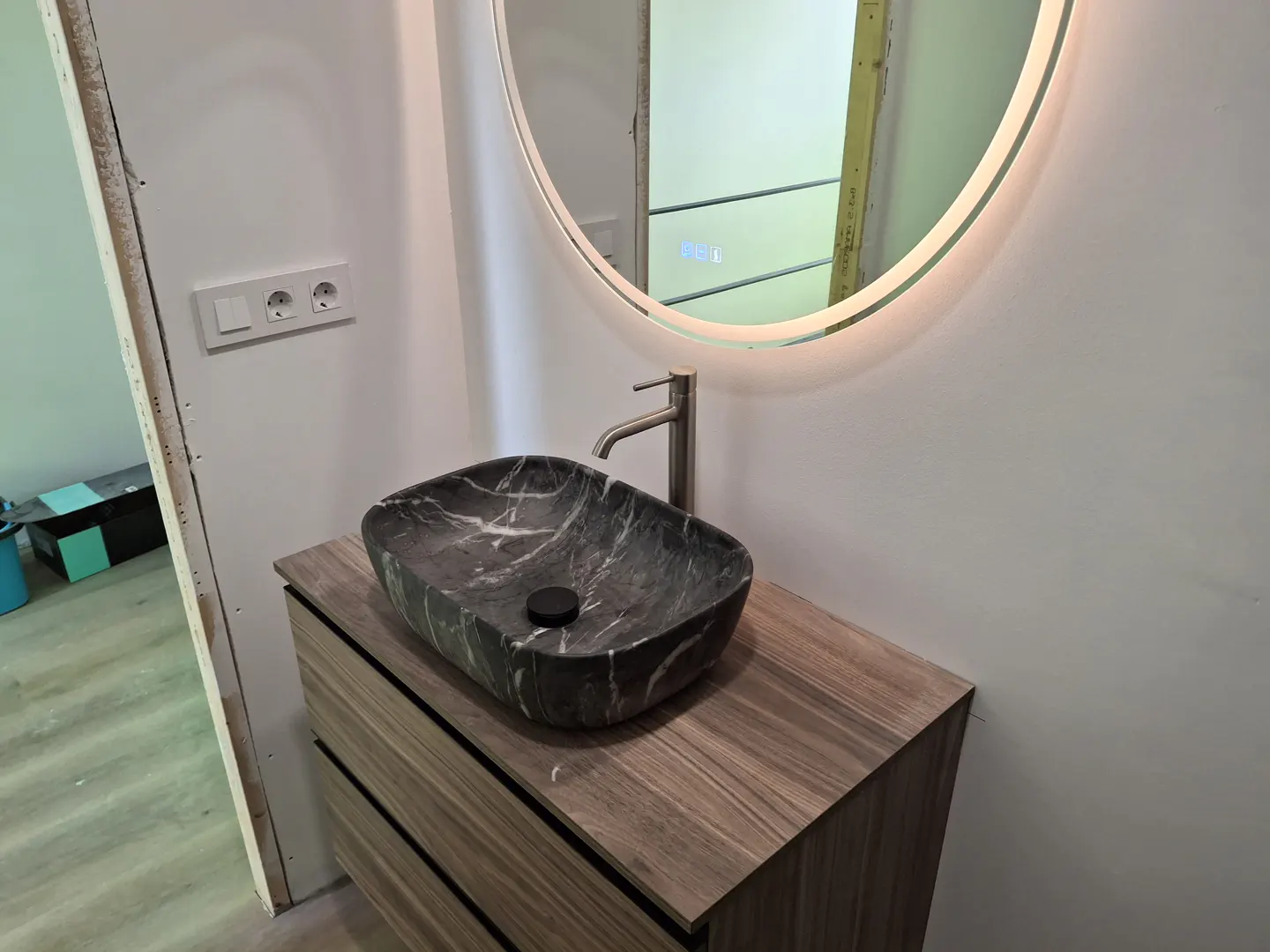 Bathroom vanity with a black marble sink, silver faucet, and round LED mirror. The vanity has a wood grain finish and drawer pulls.