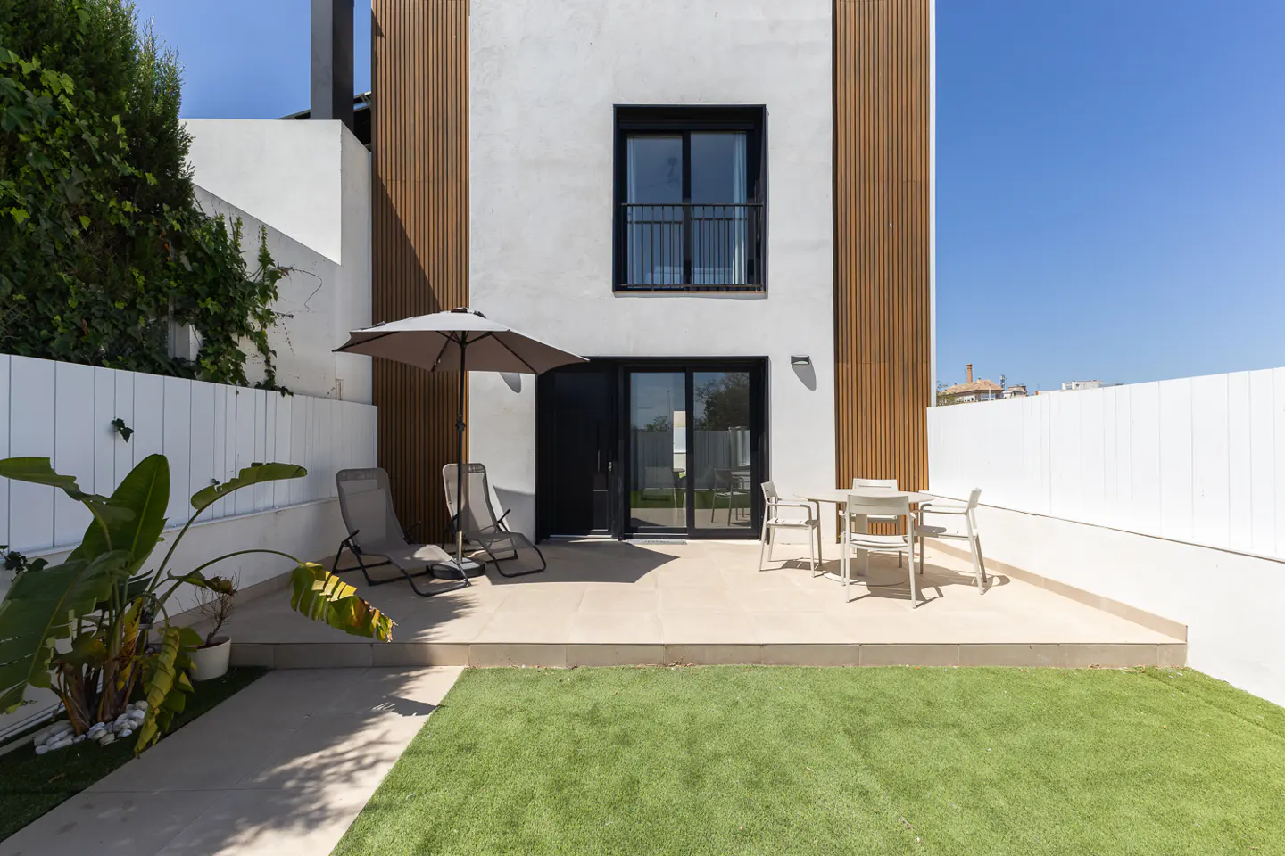 Modern home exterior with a patio, lawn, and white fence. Patio furniture includes chairs, a table, and an umbrella. The building has white walls and wood paneling.