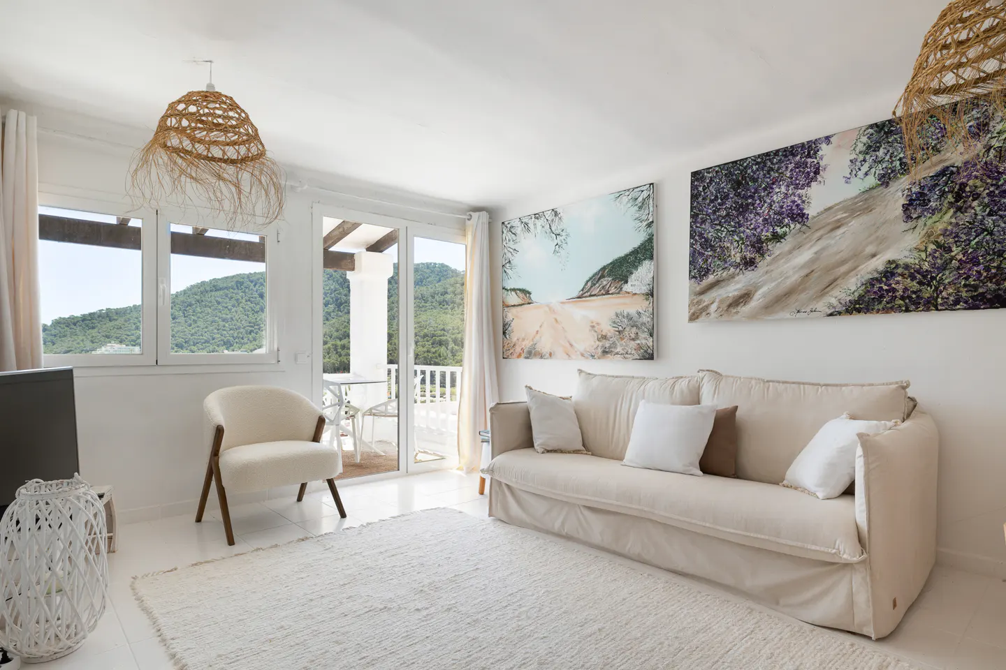 Bright living room with white walls, a beige sofa, and a white rug. Two landscape paintings hang above the sofa. A balcony overlooks green hills.