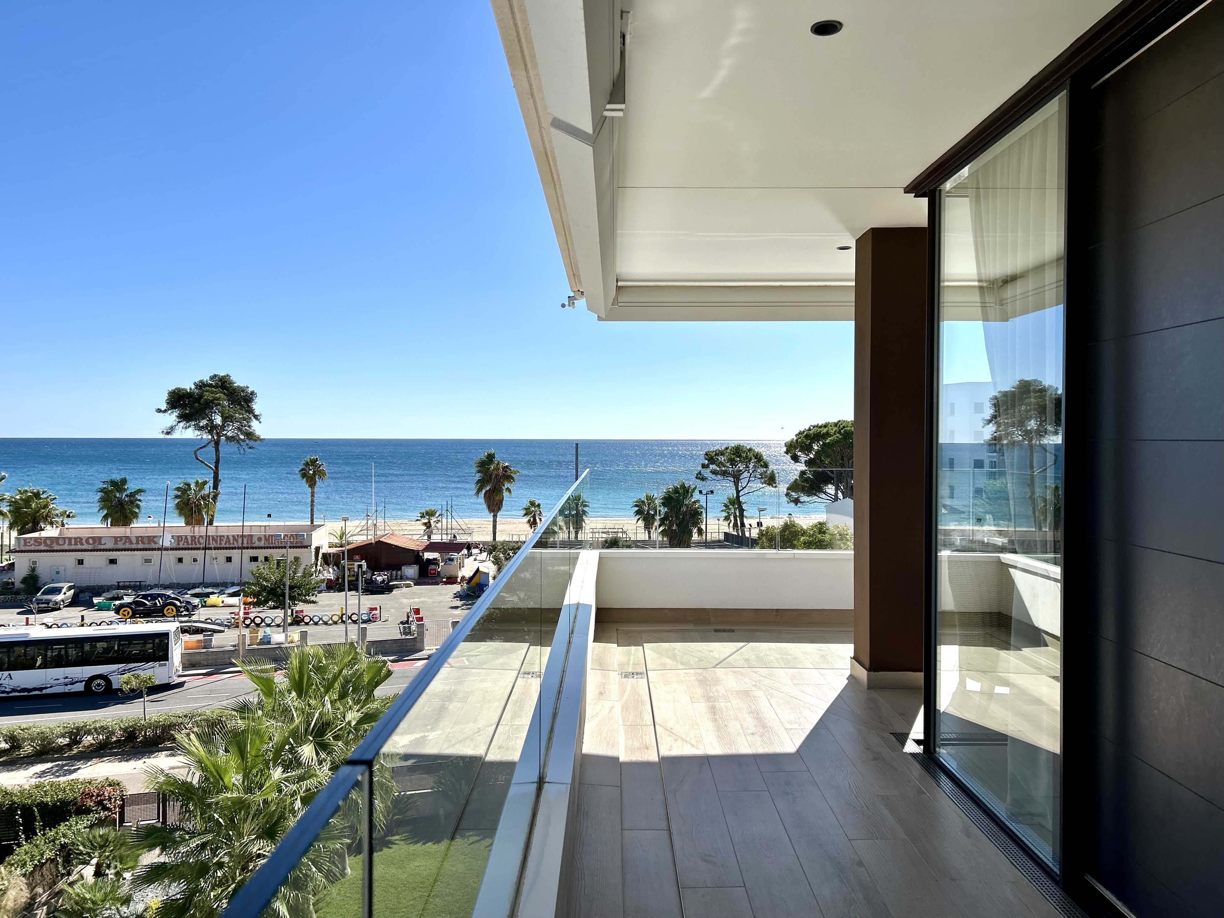 Balcony view of a beach with palm trees and blue ocean under a clear sky. Glass railing and sliding door visible.