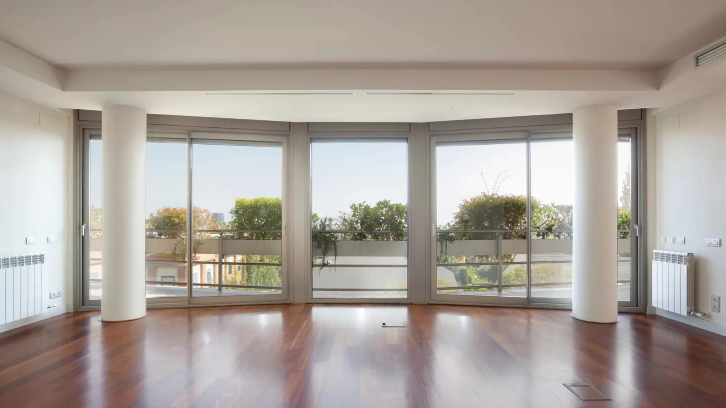 Empty room with hardwood floors, white walls, and large windows showing a balcony with greenery.