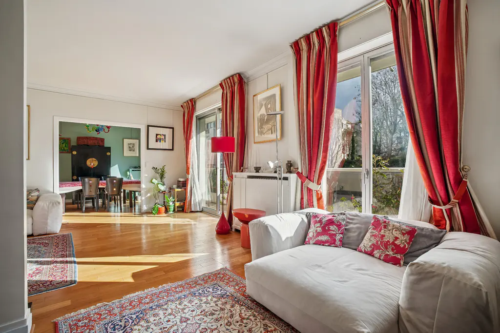 Bright living room with hardwood floors, white sofa with floral pillows, and red striped curtains framing a window. Dining room visible in background.