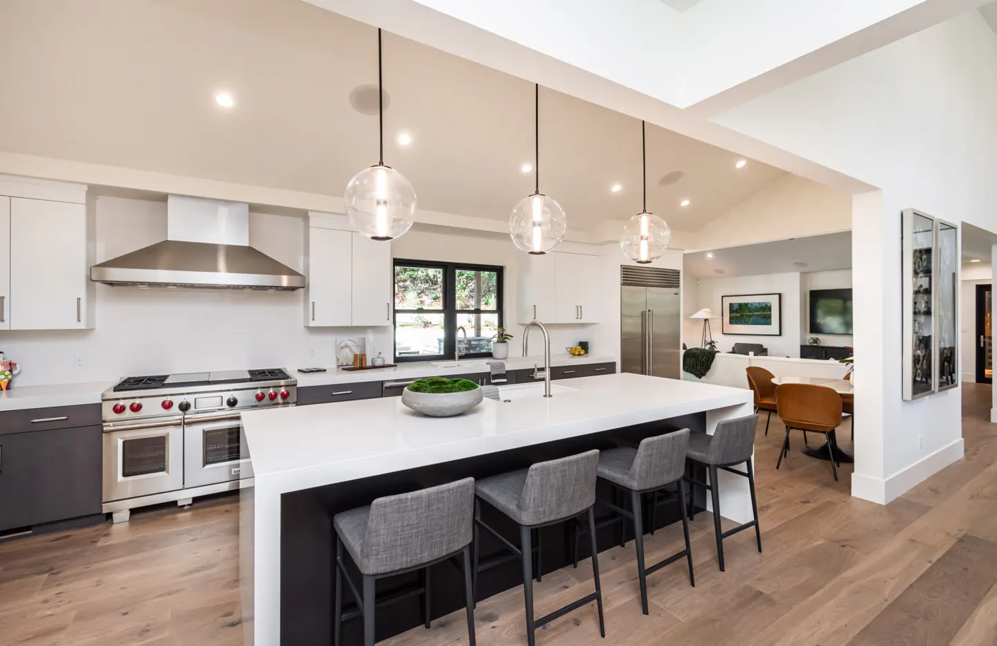 Bright, modern kitchen with white cabinets, stainless steel appliances, and a large island with gray stools. Three globe pendant lights hang above.