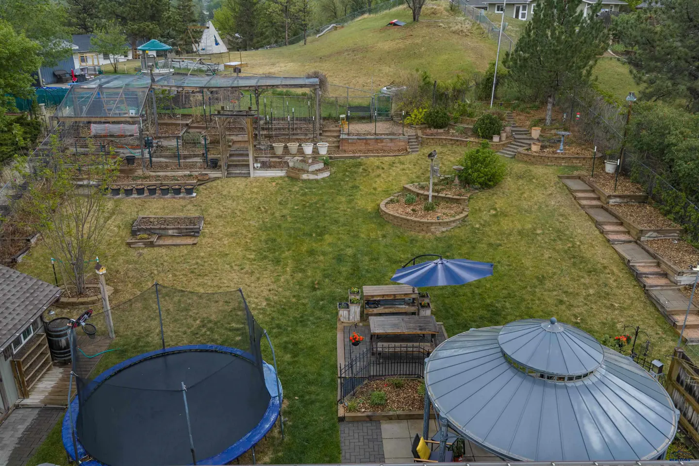 High-angle view of a backyard with a trampoline, a gazebo, a patio dining set with a blue umbrella, and terraced gardens.