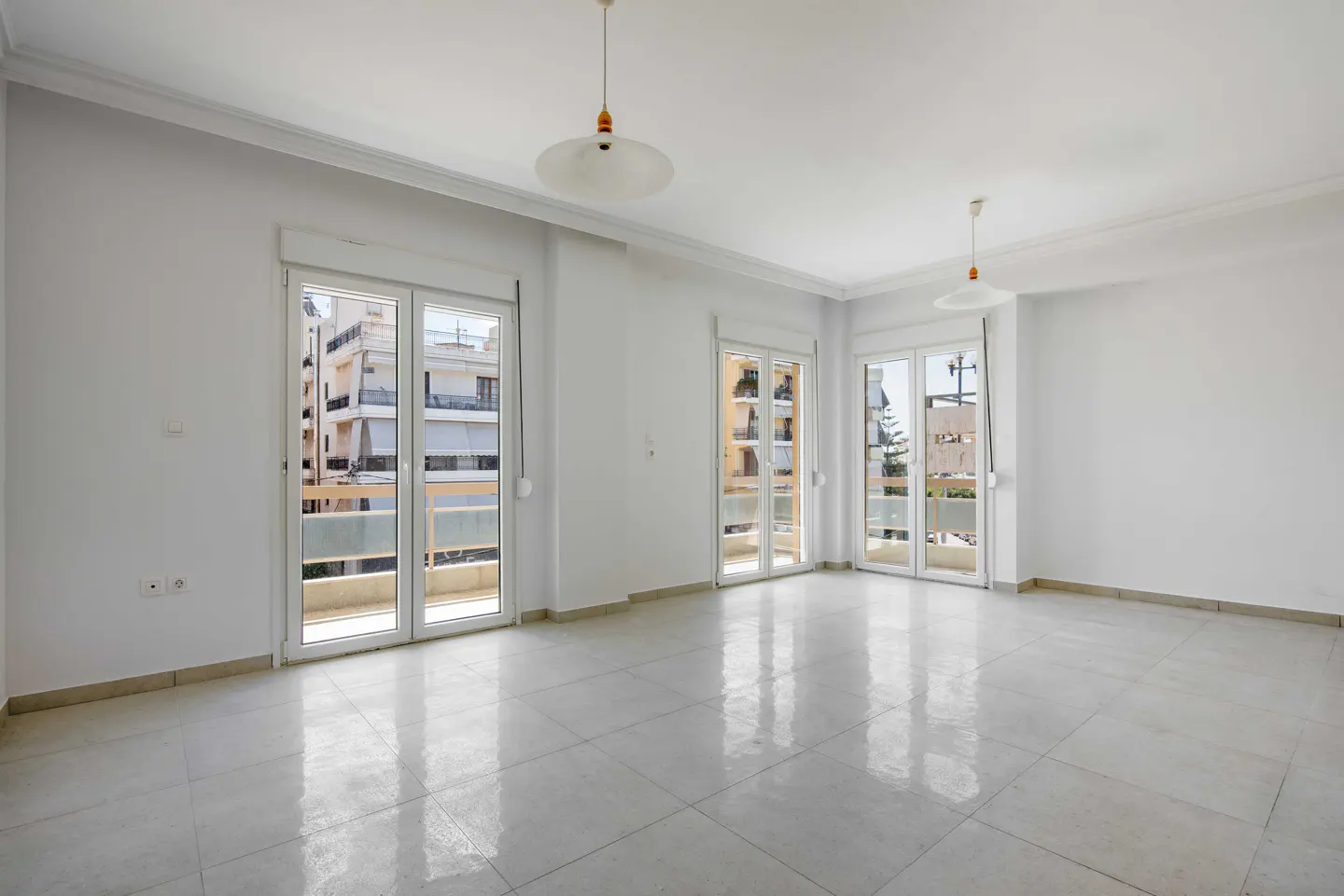 Bright, empty room with white walls, tile floor, and three sets of glass doors to balconies. Two pendant lights hang from the ceiling.