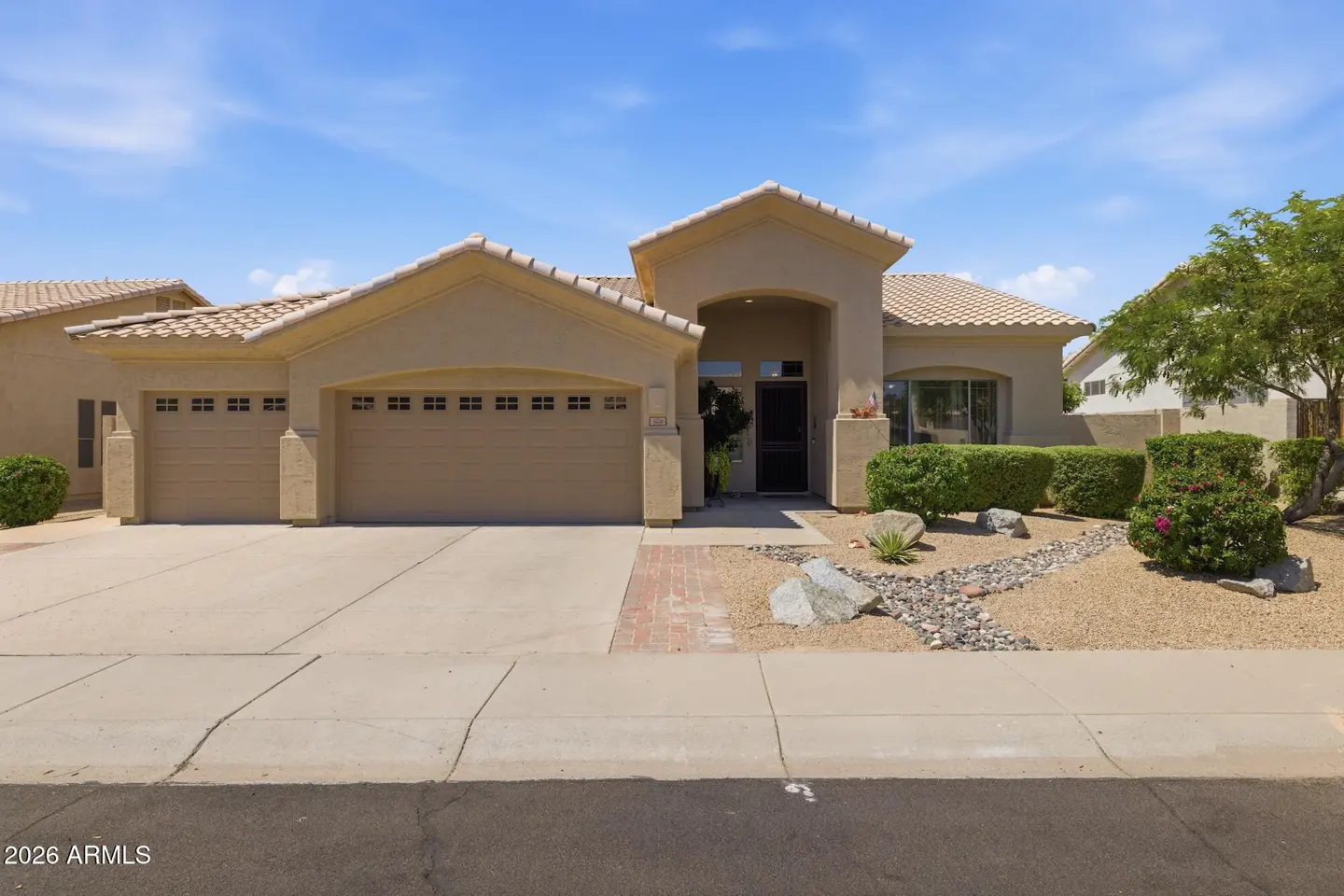 Single-story tan stucco home with a three-car garage, desert landscaping, and a blue sky background.
