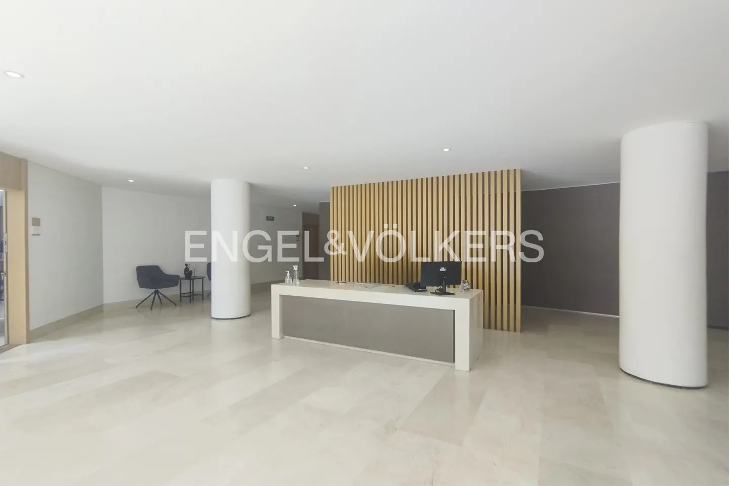Modern real estate office lobby with a white reception desk, computer, and wood slat wall. Marble floors and white columns.