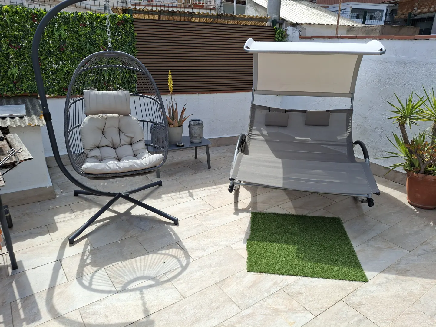 A sunny rooftop terrace featuring a hanging egg chair, a shaded lounge swing, potted plants, and a small side table.